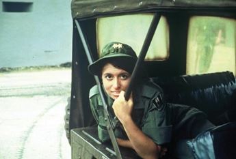 A woman in military attire looks at the camera from a vehicle.