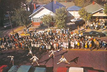 A yearbook photo of a pep rally on a Dodge City street. Cheerleaders jump in the foreground while a large crowd looks on.