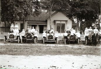 historic photo of people sitting on parked cars in front of a home