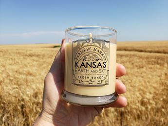 A hand holds a candle in a glass jar in front of a blue sky and field of wheat.