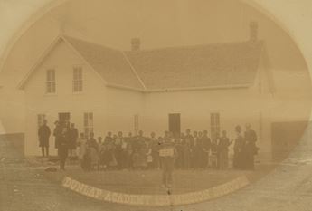 Students and teachers stand outside a schoolhouse. The words "Dunlap Academy and Mission School" and across the bottom of the photo. Sepia tone.