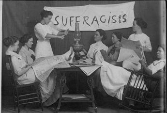 Group of women dressed in white meet around a table with a banner reading "Sufferagists" in the background.