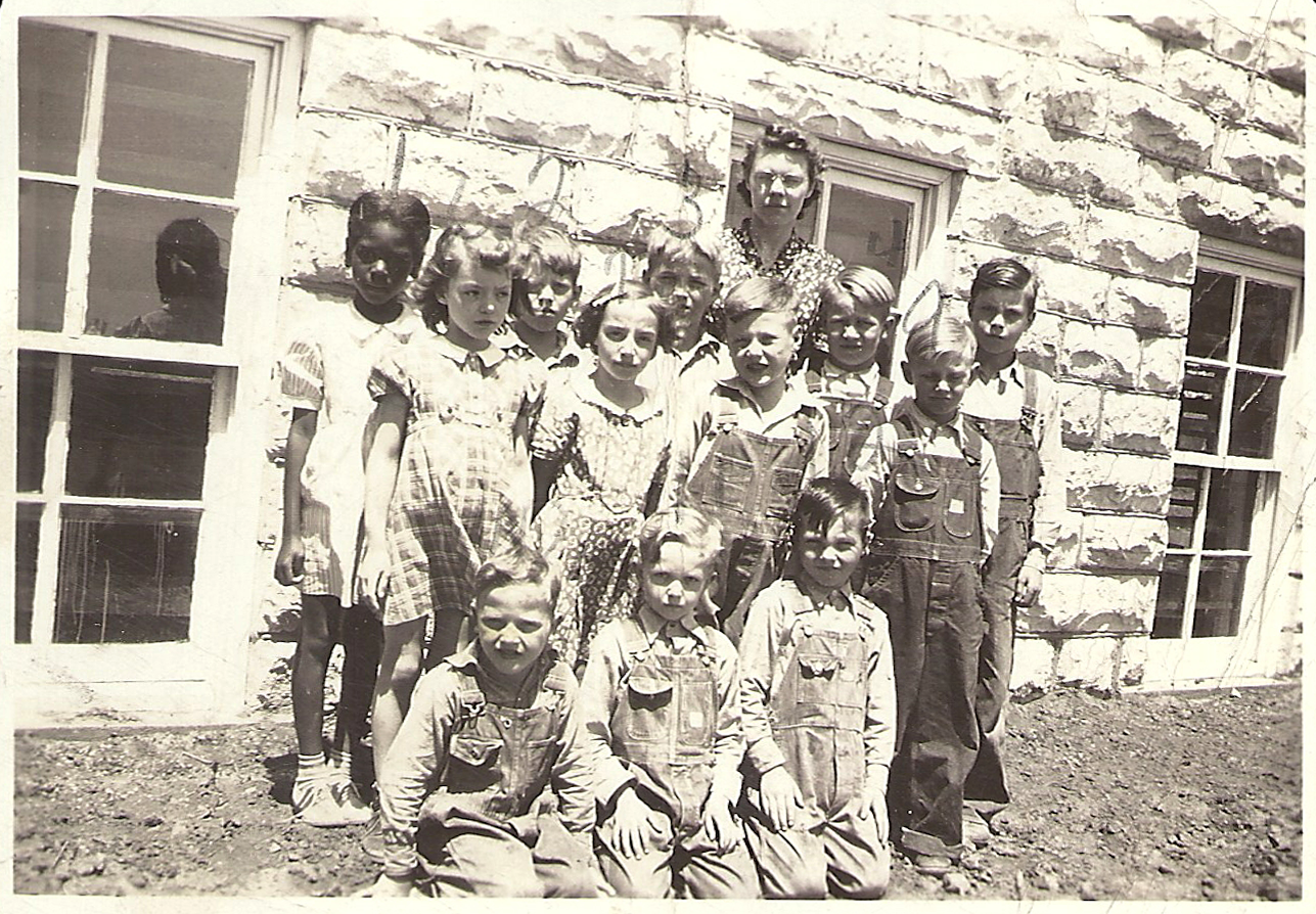 A teacher stands with students in front of a schoolhouse. Black and white photograph.