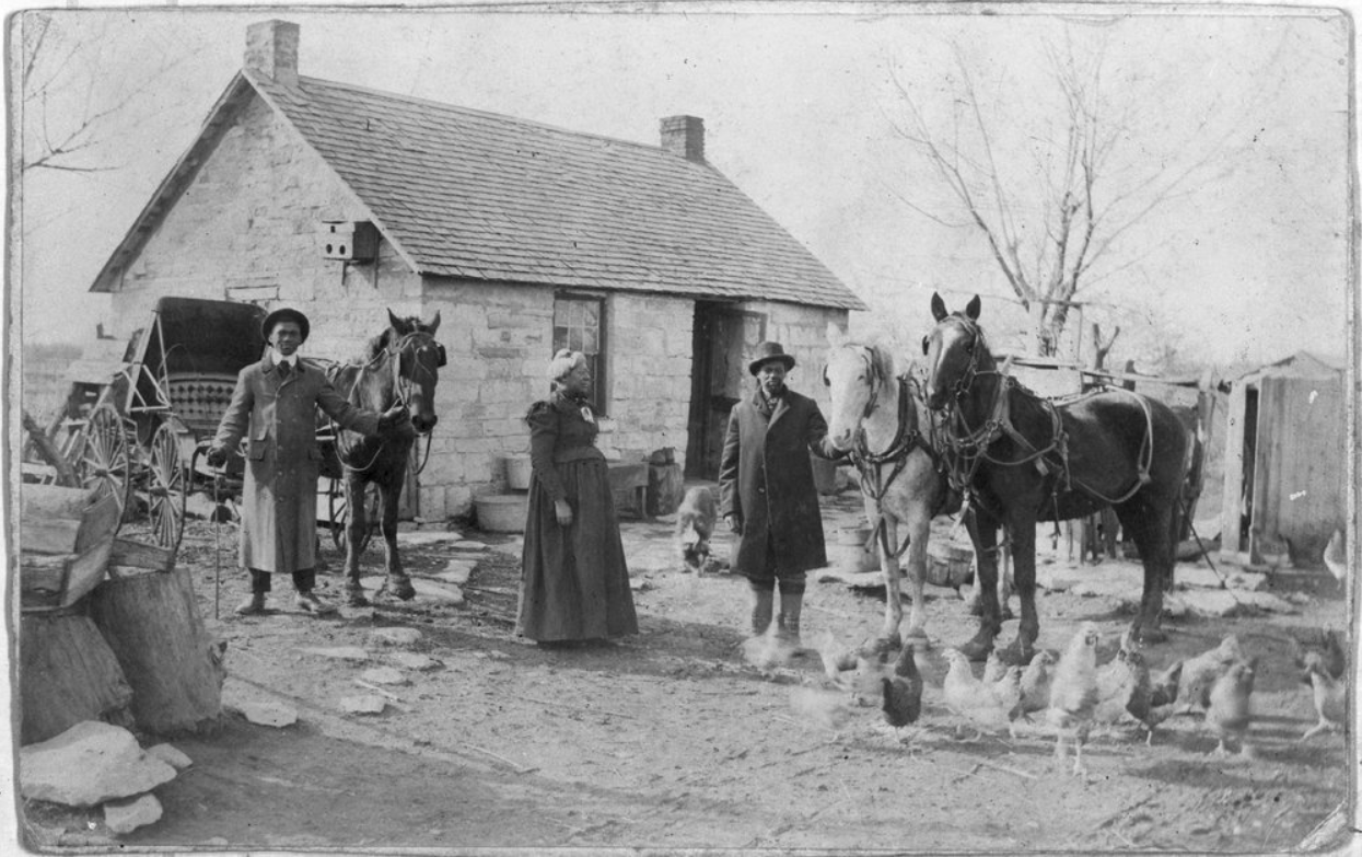 Three people stand with three horses and chickens in front of a house. Black and white photograph.