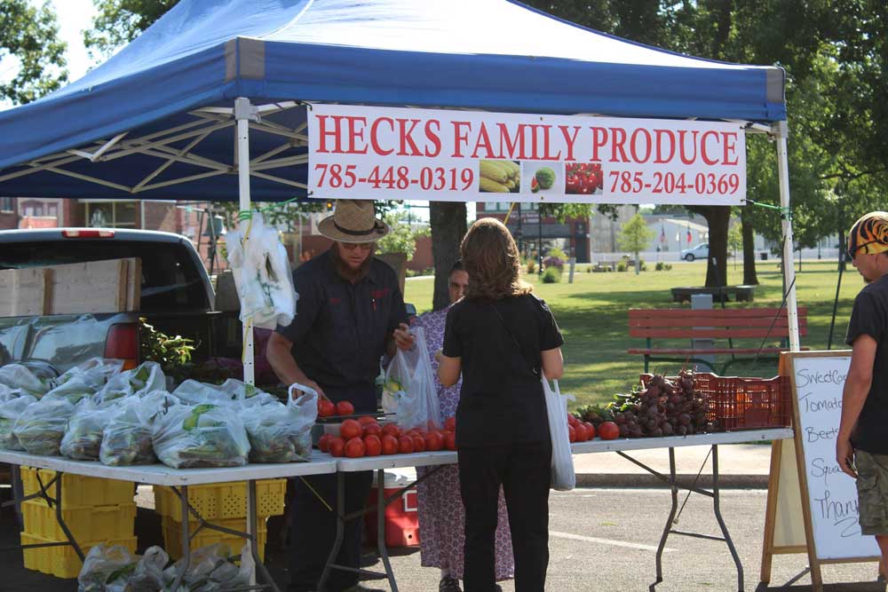 Photo of a produce stand under a tent at the Iola Farmers' Market. The banner says Hecks Family Produce.